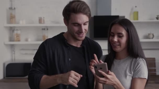 Het echtpaar in hun keuken kijken samen naar de smartphone en veel plezier met praten, ze gesticuleren en glimlachen — Stockvideo