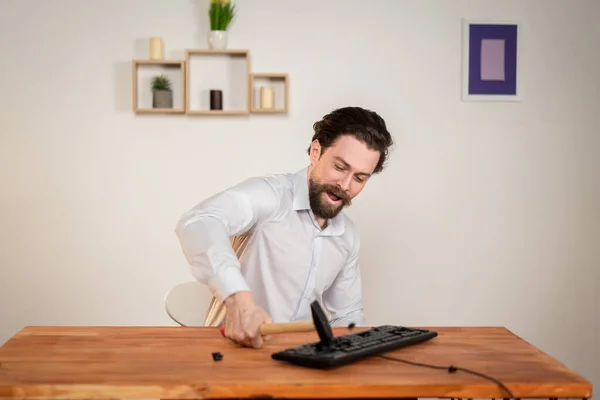 Trabajador de oficina agresivo, hombre enojado con barba destruir su ordenador, con martillo en las manos destruir el teclado. — Foto de Stock