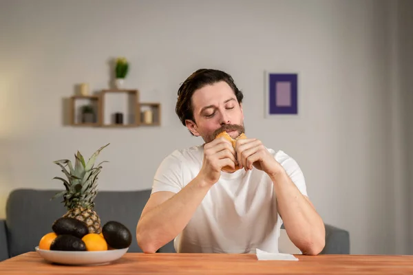 Um jovem senta-se à mesa e fala de comida, pega no hambúrguer e come-o. — Fotografia de Stock