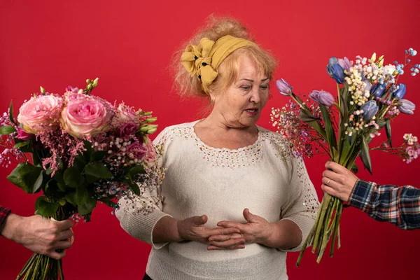 Yaşlı bir kadın buketi seçer, iki farklı buket çiçeğe bakar ve en iyisinin ne olduğunu anlamaya çalışır, bayram konsepti, hediye seçenekleri