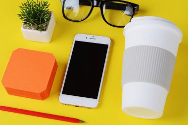 Plastik bardak kahve, kalem, telefon ve gözlük ile boş not kağıdı. Sarı arka plan