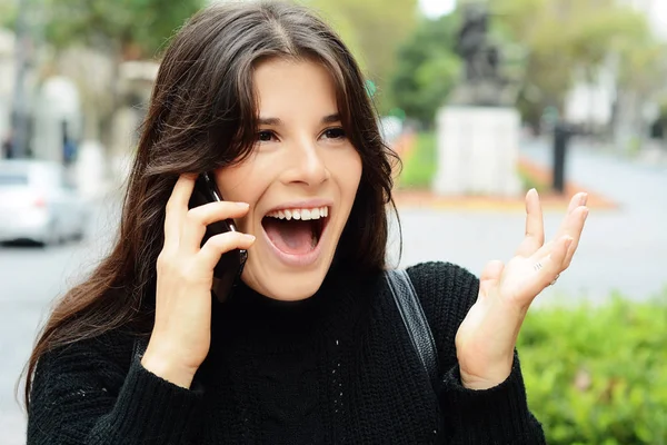 Portrait of a surprised woman with open mouth while talking on smart phone. Outdoors city urban background.