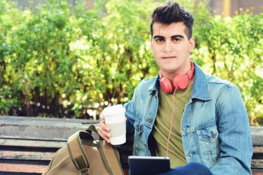 Tablet ile bir bankta oturmuş ve içme kahve parkta genç adam. Açık havada