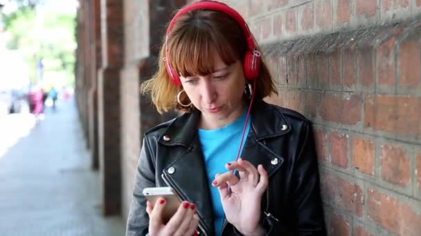 一个年轻的红头发女孩在砖墙上用智能手机和耳机选择音乐的肖像 — 图库视频影像