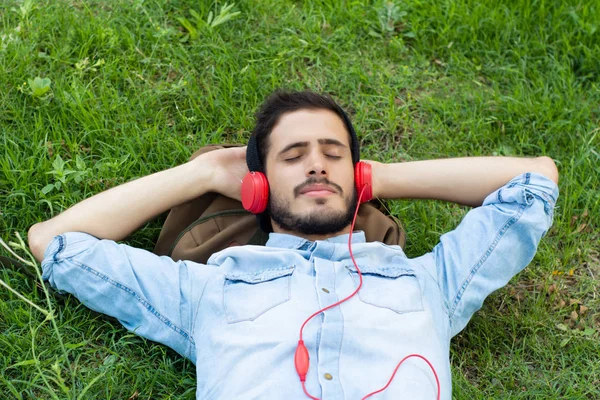 Genç adam kulaklık ile yeşil çimenlerin üzerinde rahatlatıcı. Boş zaman kavramı.