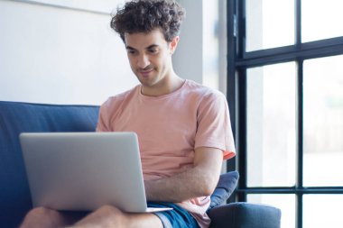 Genç adam elinde dizüstü bilgisayarla kanepede dinleniyor. 