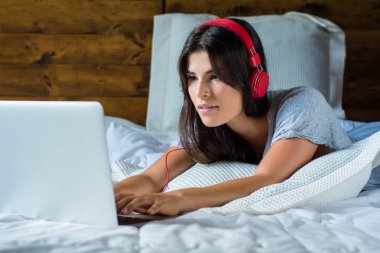 Genç kadın yatakta laptop ve kulaklık kullanarak.