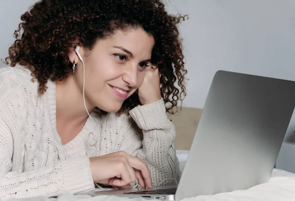 Young woman using laptop and headphones in bed.