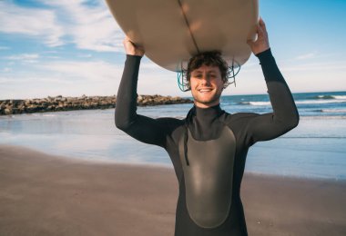 Plajda sörf tahtasını kaldırıp siyah bir sörf kıyafeti giyen genç sörfçünün portresi. Spor ve su sporları konsepti.