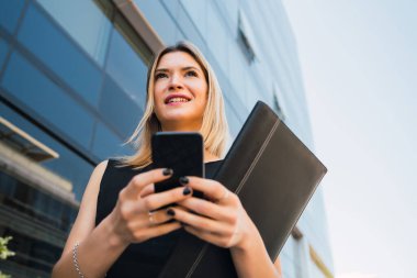 Ofis binalarının önünde beklerken cep telefonunu kullanan genç bir iş kadınının portresi. İş ve başarı kavramı.