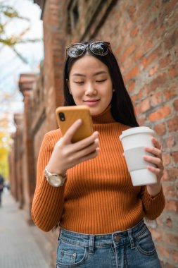 Sokakta elinde kahve tutarken cep telefonunu kullanan Asyalı genç bir kadının portresi. Kentsel ve iletişim konsepti.