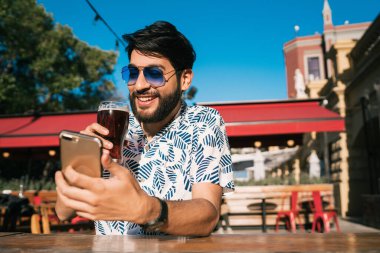 Genç yakışıklı adam yaz havasının tadını çıkarıyor ve dışarıda bira içerken cep telefonunu kullanıyor. İletişim konsepti.
