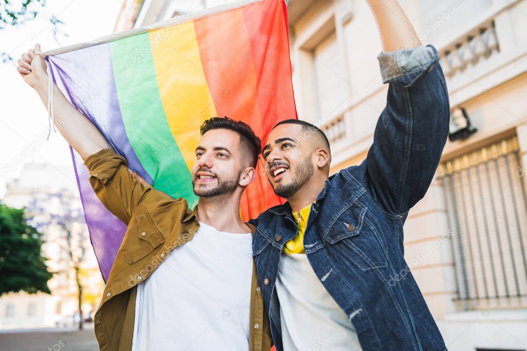 Portrait of young gay couple embracing and showing their love with rainbow flag at the street. LGBT and love concept