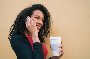 Sarı arka planda bir fincan kahve tutarken telefonla konuşan genç afro kadın portresi. İletişim konsepti.