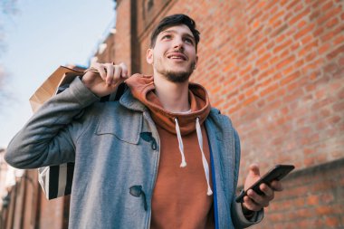 Sokakta yürüyen, alışveriş torbaları tutan ve cep telefonunu kullanan yakışıklı bir adamın portresi. Kentsel, alışveriş konsepti.