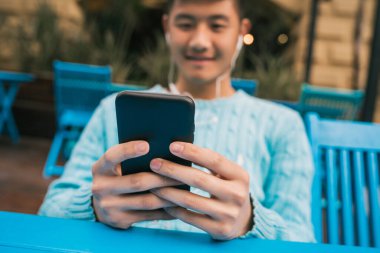 Bir kafede otururken cep telefonuyla kulaklık takan Asyalı genç bir adamın portresi. İletişim konsepti.