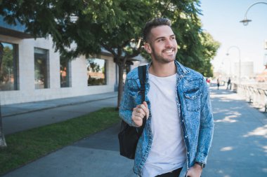 Omuzlarında sırt çantasıyla sokakta yürüyen çekici bir gencin portresi. Şehir konsepti.