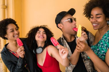 Çok ırklı arkadaş grubunun portresi, sarı arka planda dondurma yerken eğleniyor ve eğleniyor..