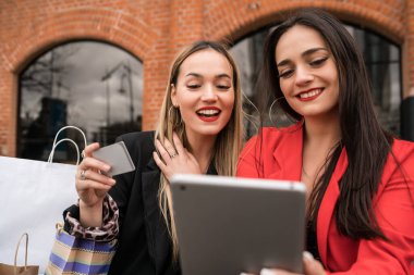 Açık havada otururken kredi kartı ve dijital tabletle alışveriş yapan iki genç arkadaşın portresi. Arkadaşlık ve yaşam tarzı kavramı.
