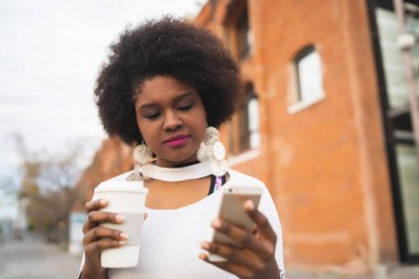 Sokakta elinde bir fincan kahve tutarken cep telefonunu kullanan Afro Latin kadının portresi. Kentsel ve iletişim konsepti.