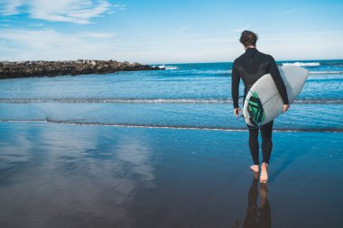 Genç sörfçü siyah sörf kıyafetiyle sörf tahtasıyla suya giriyor. Spor ve su sporları konsepti.