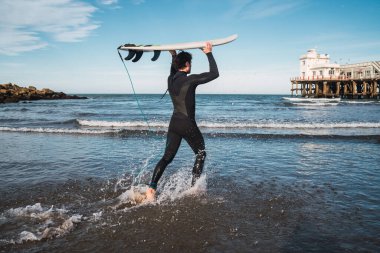 Genç sörfçü siyah sörf kıyafetiyle sörf tahtasıyla suya giriyor. Spor ve su sporları konsepti.