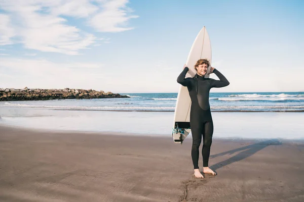 Portrait of young surfer at the beach holding up his surfboard and wearing a black surfing suit. Sport and water sport concept.