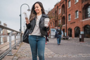Bir fincan kahveyle yürürken cep telefonunu kullanan genç bir kadının portresi. Kentsel ve iletişim konsepti.
