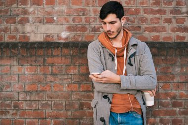 Kulaklıklı genç bir adamın portresi ve tuğla duvara karşı cep telefonu kullanması. Açık havada. Şehir konsepti.