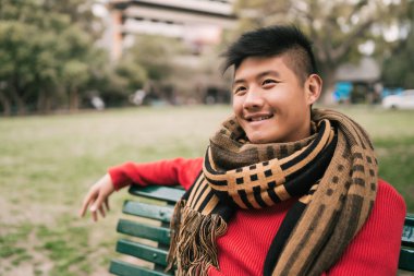 Genç Asyalı bir adamın portresi, kışlık giysiler giymiş ve rahatlamış bir şekilde parkta bir bankta oturuyor.. 