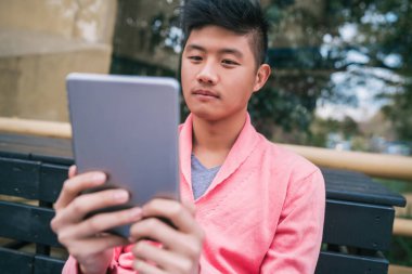 Dışarıdaki bankta otururken dijital tabletini kullanan genç Asyalı adamın portresi. Teknoloji konsepti.