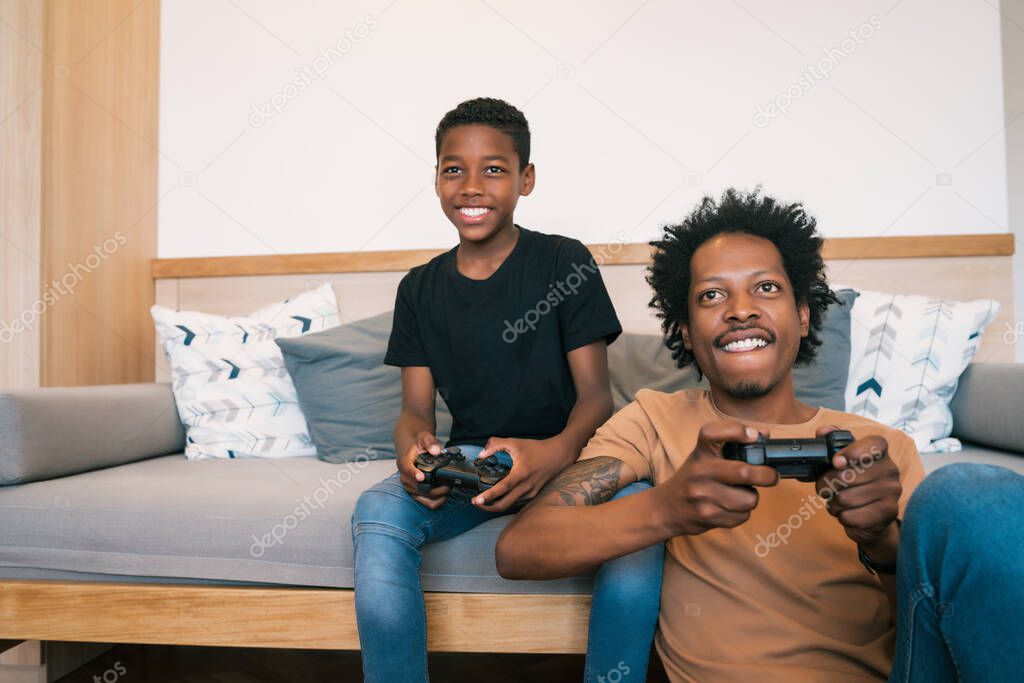 Portrait of happy African American father and son sitting in sofa couch and playing console video games together at home. Family and technology concept.