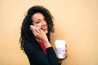 Sarı arka planda bir fincan kahve tutarken telefonla konuşan genç afro kadın portresi. İletişim konsepti.