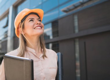 Profesyonel mimar kadın portresi sarı kask takıyor ve açık havada modern binalara bakıyor. Mühendis ve mimar kavramı.