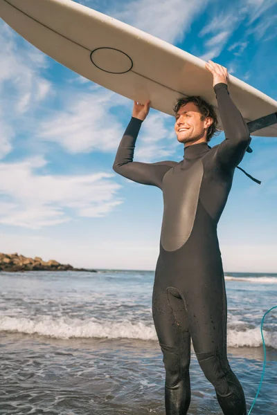 Portrait of young surfer at the beach holding up his surfboard and wearing a black surfing suit. Sport and water sport concept.