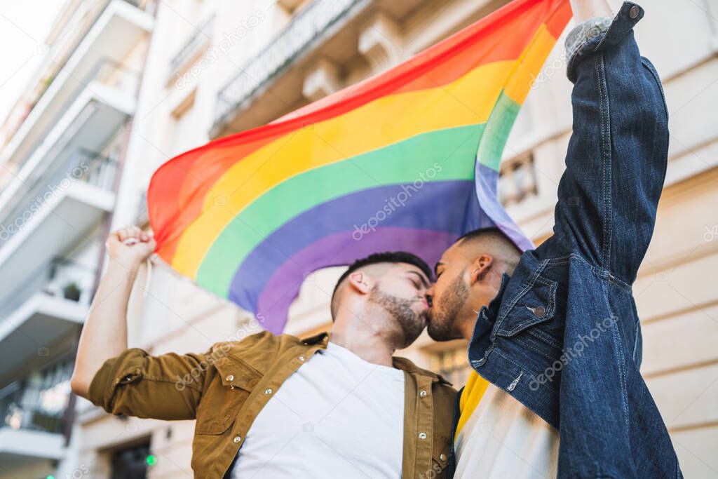 Portrait of young gay couple embracing and showing their love with rainbow flag at the street. LGBT and love concept