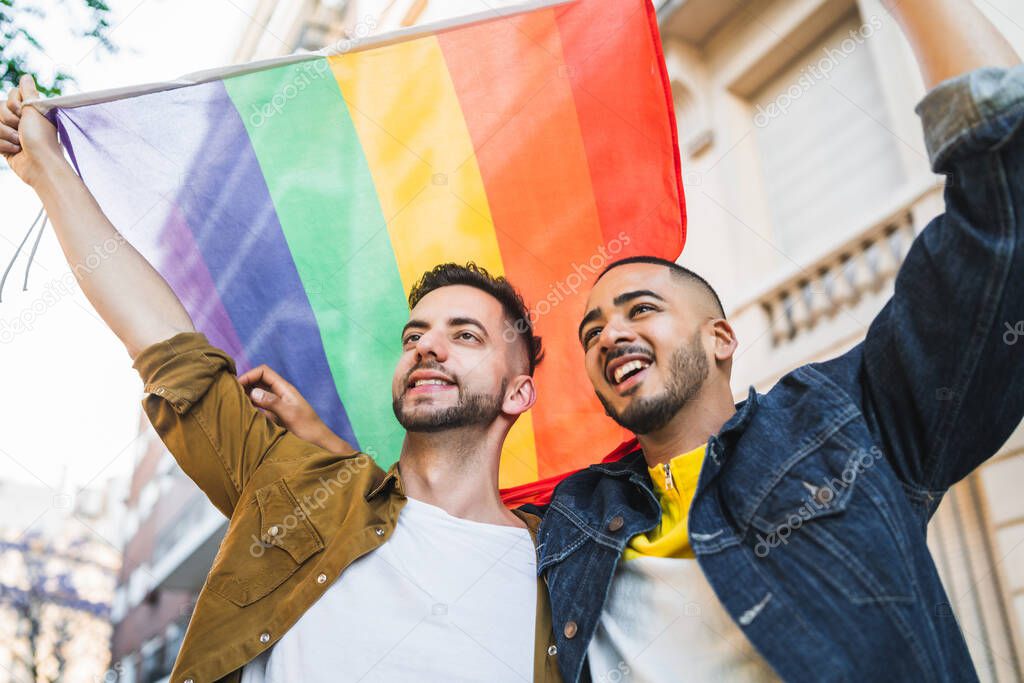 Portrait of young gay couple embracing and showing their love with rainbow flag at the street. LGBT and love concept