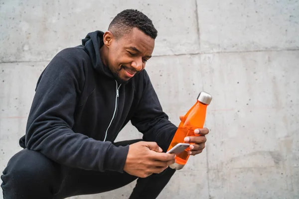 Athletic man using his mobile phone and holding a bottle of water after training against grey background. Sport and health lifestyle.