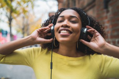 Afro-Amerikan kadın portresi gülümseyen ve sokakta kulaklıkla müzik dinleyen. Açık havada..