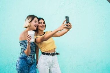 Lezbiyen bir çiftin portresi. Açık mavi arka planda cep telefonuyla selfie çekiyorlar. LGBT kavramı.