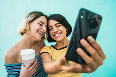 Lezbiyen bir çiftin portresi. Açık mavi arka planda cep telefonuyla selfie çekiyorlar. LGBT kavramı.