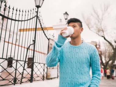 Sokakta yürürken bir fincan kahve içen genç Asyalı adamın portresi. Şehir konsepti.