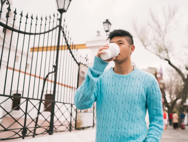 Sokakta yürürken bir fincan kahve içen genç Asyalı adamın portresi. Şehir konsepti.