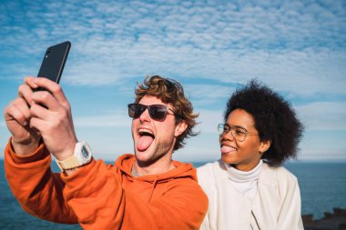 İki genç arkadaşın birlikte iyi vakit geçirdikleri ve deniz kenarında akıllı telefonlarla selfie çektikleri portre..