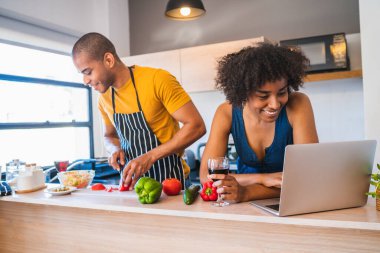 Evde yemek pişirirken dizüstü bilgisayar kullanan genç bir Latin çiftin portresi. İlişki, aşçılık ve yaşam tarzı kavramı.
