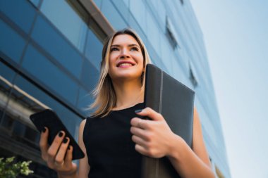 Ofis binalarının önünde beklerken cep telefonunu kullanan genç bir iş kadınının portresi. İş ve başarı kavramı.
