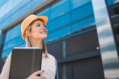 Profesyonel mimar kadın portresi sarı kask takıyor ve açık havada modern binalara bakıyor. Mühendis ve mimar kavramı.