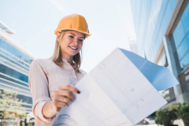 Profesyonel mimar portresi sarı kask takıyor ve modern binanın dışındaki mavi izlere bakıyor. Mühendis ve mimar kavramı.