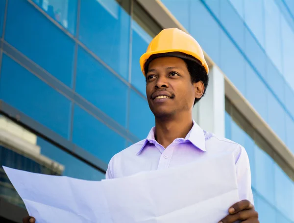 Portrait of professional architect in helmet looking at blue prints outside modern building. Engineer and architect concept.