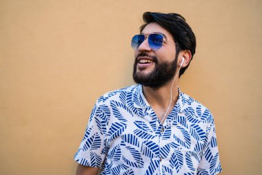 Yazlık kıyafetler giyen ve sarı arka planda kulaklıkla müzik dinleyen genç Latin adamın portresi..